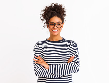 Positive ethnic woman in striped garment and trendy glasses smiling for camera and crossing arms against gray backgroundの写真素材