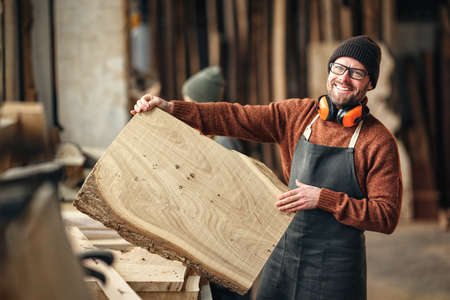 Positive adult bearded craftsman in apron showing large wooden board while standing near workbench in carpentry workshopの写真素材