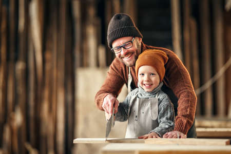 Smiling little boy learning to cut wooden plank with saw with help of father while spending time together in joinery workshopの写真素材