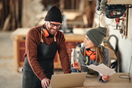 Positive man and woman in aprons and with protective headphones standing near table with laptop and discussing ideas for project while working together in professional carpentry workshopの写真素材