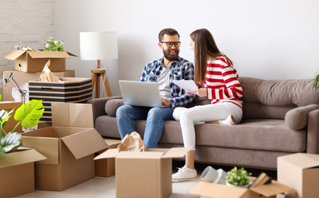 Happy young family couple with laptop and papers sitting on sofa in room with packed boxes and...