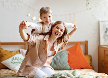 Excited mother laughing and carrying cheerful boy on back while kneeling on bed in cozy bedroom in...