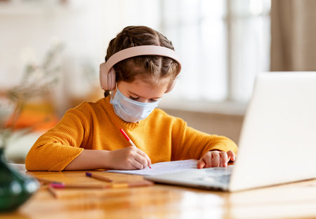 Focused serious elementary little girl in medical mask and headphones making notes in notepad while communicating with teacher through video chat app during online education at homeの写真素材