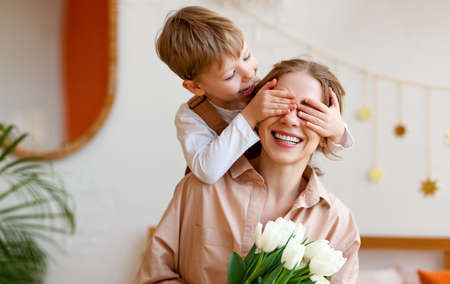 tender son closes his mother's eyes, making her a surprise and gives her a tulips, congratulating her on mother's day during celebration at homeの写真素材