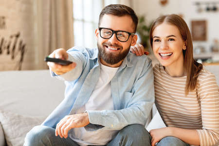 Delighted adult couple: man and woman smiling and looking at camera while sitting on sofa and changing channels on TV with remote controlの写真素材