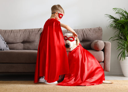 Full body side view of little boy and cute Labrador retriever dog dressed in red superhero cloaks and glasses playing together at homeの写真素材
