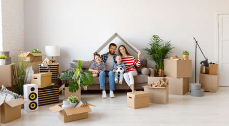 Cheerful parents and kids holding house roof over head and smiling for camera while sitting on couch and belongings in new flatの写真素材