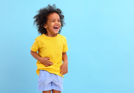 Delighted little black kid with Afro hair in casual clothes laughing and looking away against blue backgroundの写真素材