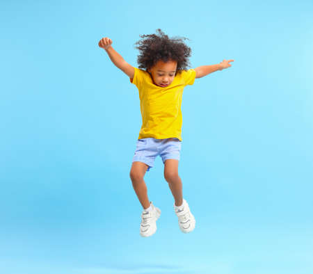 Full length of energetic little ethnic boy with Afro hairstyle in stylish casual outfit jumping with raised arms and looking down against blue backgroundの写真素材