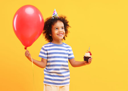 Cheerful cute little African American boy with curly hair in casual clothes smiling and looking away while standing against yellow background with red balloon and birthday cupcake in handsの写真素材