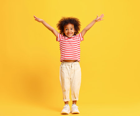 Full body of excited little African American child with curly hair in stylish outfit standing...