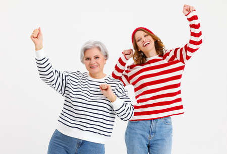 Positive middle aged gray haired woman and teenage daughter wearing colorful striped shirts and jeans having fun and dancing with arms raised on white backgroundの写真素材