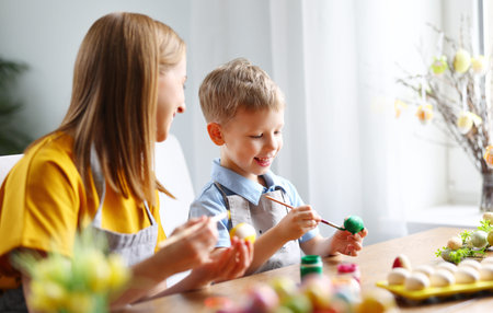 Happy family: mother and son sitting at table at home and painting eggs with gouache while preparing...