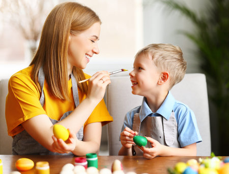 Playful mother painting nose of adorable boy sitting at table while preparing eggs for celebration...