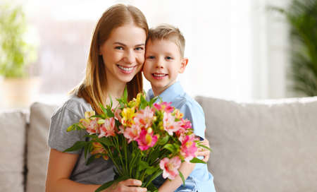 Cheerful son embracing mother sitting on sofa with bouquet of Alstroemeria flowers on Mothers Dayの写真素材