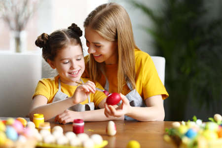 Happy family: mother and daughter sitting at table at home and painting eggs with gouache while preparing for Easter holidayの写真素材
