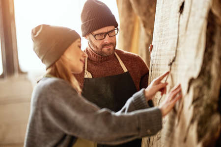 couple of colleagues male and female artisan masters in aprons standing near large lumber plank while working together in carpentry workshopの写真素材