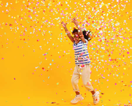 Full body of joyful little black child with Afro hair in stylish clothes laughing and jumping while trying to catch colorful confetti on birthday against yellow backgroundの素材 [FY310166224660]