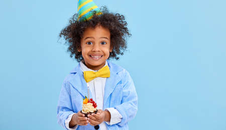 Adorable happy African American little boy in bow tie and party hat smiling and looking at camera while standing against blue background with yummy birthday cupcake in handsの写真素材