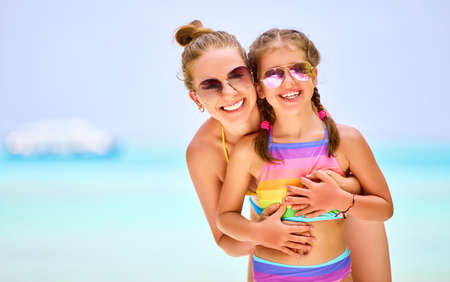 Happy woman and charming girl in colorful swimwear and sunglasses looking at camera happily on blurred background of oceanの写真素材