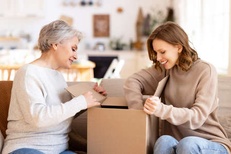 Positive curious young woman with middle aged mother sitting on sofa and unpacking cardboard box with ordered purchases while spending time together at homeの写真素材