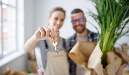 Content young couple with keys from new apartment embracing while standing in room with various...