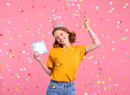 Happy young female in casual clothes and party hat with gift box in hand dancing under colorful confetti during birthday celebration against pink backgroundの写真素材
