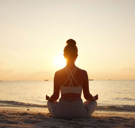 Back view of female sitting in Padmasana while meditating alone on seashore against sky in sundown lightの写真素材