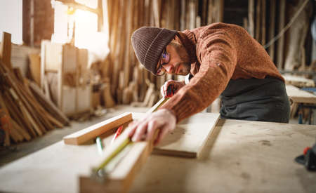 Focused adult male woodworker in apron and knitted gat cutting wooden plank while working in carpentry workshopの写真素材