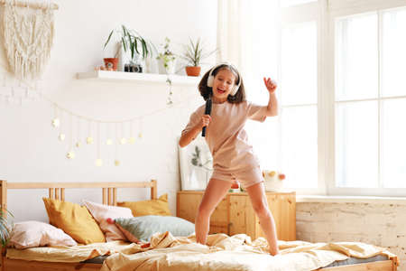 Little star. Cute happy little girl wearing pajama with hairbrush as mic pretending to be famous singer while standing on bed at home, adorable preschool child singing and dancing in bedroomの写真素材