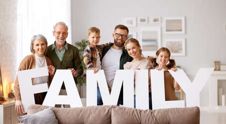 Housing and mortgage concept. Big happy family of six people grandparents, mother and father with little kids holding word FAMILY smiling at camera while sitting on sofa in the living room in new homeの素材 [FY310167762725]