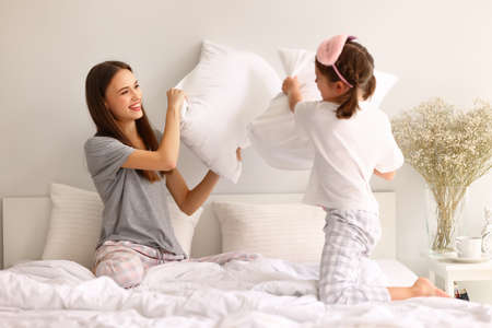 Cheerful family: young mother and daughter hitting each other with pillows while playing on bed in weekend morning at homeの写真素材