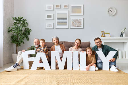 Housing and mortgage concept. Big happy family of six people grandparents, mother and father with little kids holding word FAMILY smiling at camera while sitting on floor never sofa in the living room in new homeの写真素材