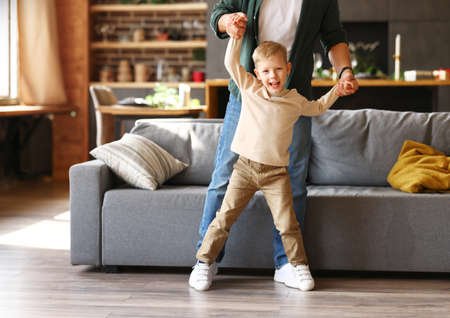 Full length shot of happy little kid boy dancing with dad in living room, holding fathers hands and smiling at camera. Happy family daddy and child playing together, enjoying leisure time at homeの写真素材