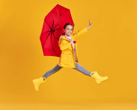 Full body side view of excited little girl in yellow raincoat and boots with red umbrella having fun and jumping above ground against yellow backgroundの写真素材