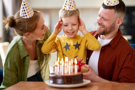 Happy little boy son going to blow candles on cake while celebrating Birthday with young loving parents, excited kid raising arms and smiling while receiving congratulations and presents from familyの写真素材