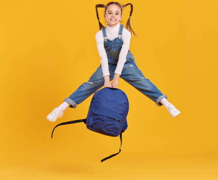 Full body of happy energetic primary schoolgirl with pigtails wearing denim jumpsuit holding backpack in hands and jumping high against yellow backgroundの写真素材