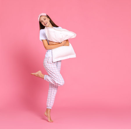 Full body delighted woman in sleepwear smiling and embracing soft pillow while standing with raised...