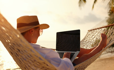 Back view of young woman, successful female freelancer using laptop while lying in hammock on the tropical beach at sunset, working remotely during summer vacation. Distance work conceptの写真素材