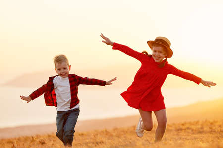 Happy smiling little children boy and girl in casual clothes running and enjoying freedom while on meadow against blurred hills and sunset sky during summer holidaysの写真素材