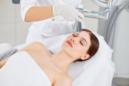 Professional cosmetologist using laser equipment for skin care treatment on face of female client in...
