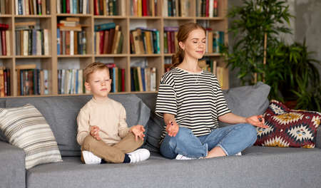 Young mom and son in casual wear meditating with closed eyes while sitting in lotus pose together on sofa in living room ar home. Calm beautiful family practicing yoga during covid19 quarantineの写真素材