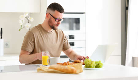 Young focused handsome man wearing glasses working remotely on laptop while sitting at table in modern kitchen at home, male freelancer healthy eating breakfast and looking at computerの写真素材