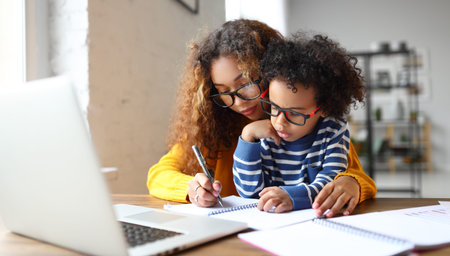 Young afro american child writing something in notebook with mother while working remotely from home...