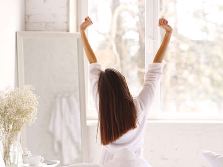 Back view of anonymous woman in sleepwear raising arms and stretching body while sitting on bed in morning in light bedroomの写真素材