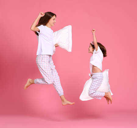 Optimistic young woman and girl with pillows raising arms and leaping up against pink backdrop in morningの写真素材