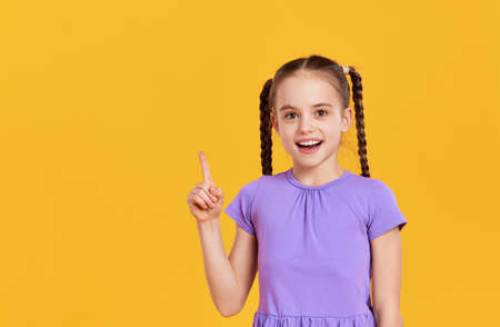 Cheerful happy little kid schoolgirl in casual clothes smiling and and looking at camera while having idea standing against yellow backgroundの写真素材
