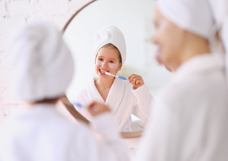 Content preteen girl in white bathrobe and towel turban with cosmetic patches under eyes brushing teeth during morning hygienic and skincare routine with momの写真素材