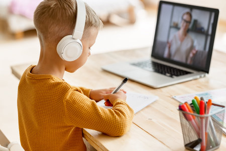 Focused child boy in headphones looks carefully at teacher while making video call during online...