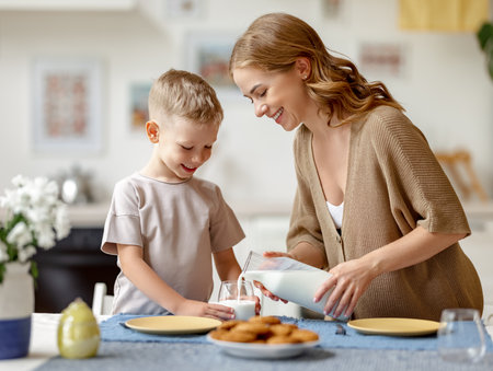 Glad woman smiling and pouring fresh milk for cheerful boy during breakfast in kitchen at homeの写真素材